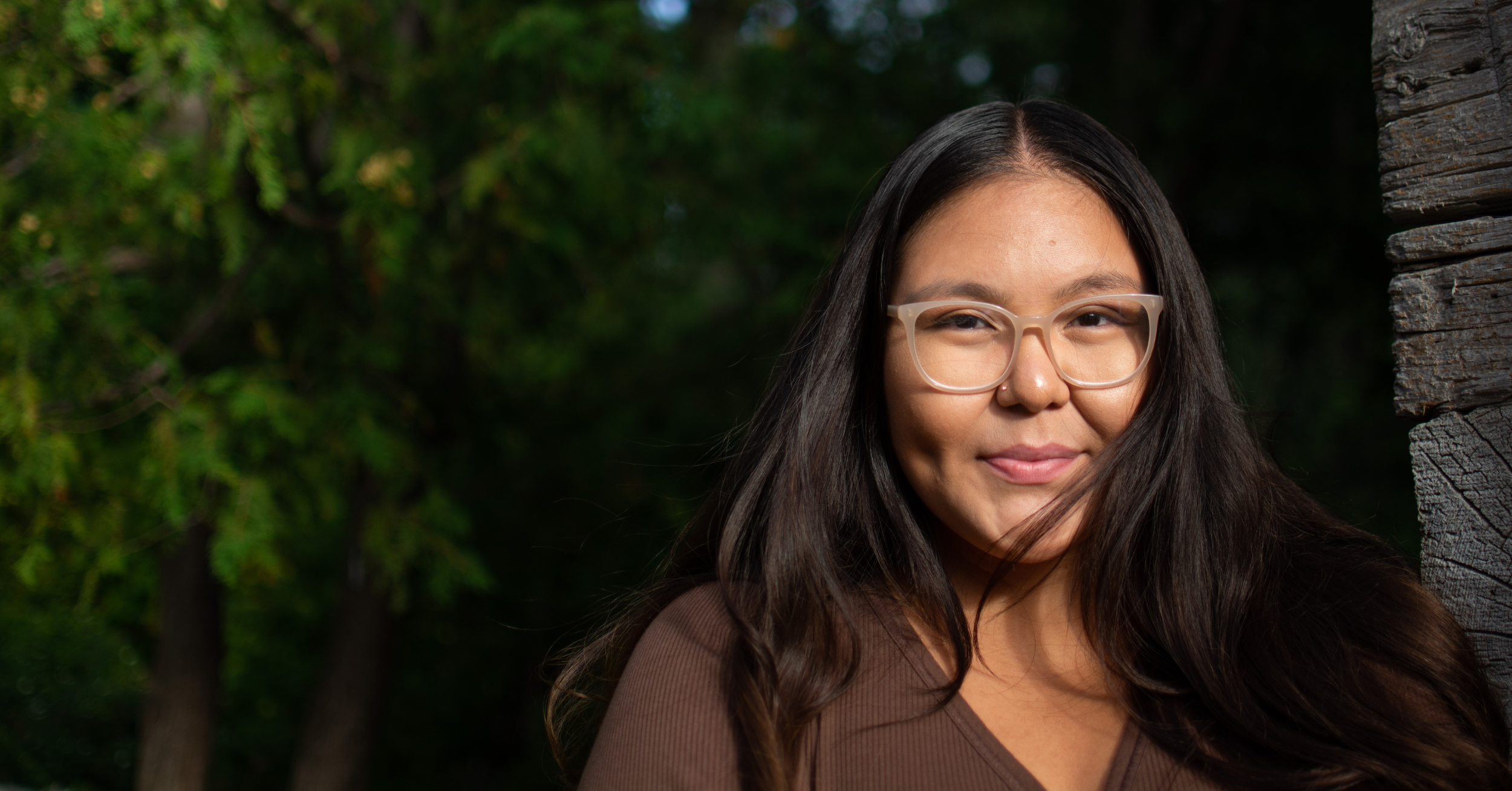 Rainingbird Daniels, a Native American woman, smiles while wearing glasses. Her long brown hair is down and flowing at both shoulders, with greenery in the background.