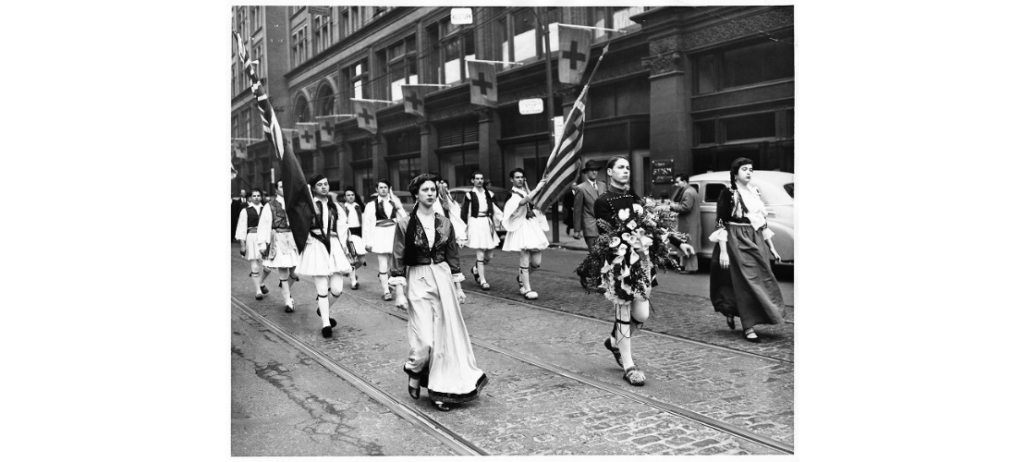 Image of group of men in traditional fustanella costume led by a young man carrying a wreath, flanked by two women in traditional Greek costume walking from St. George's Greek Orthodox Church, Bond St., west along Queen Street to the Cenotaph at City Hall (March 30, 1952). Greek Canadian History Project, Toronto Telegram Fonds, F0433, Clara Thomas Archives & Special Collections, York University Libraries. Photographer: Nelson Quarrington. ASC08802.