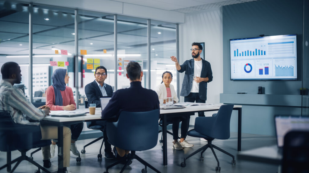 Image of a diverse modern office setting: A businessman leads a business meeting with managers, discussing and utilizing a presentation TV displaying statistics and infographics. Meanwhile, digital entrepreneurs work on an e-commerce project