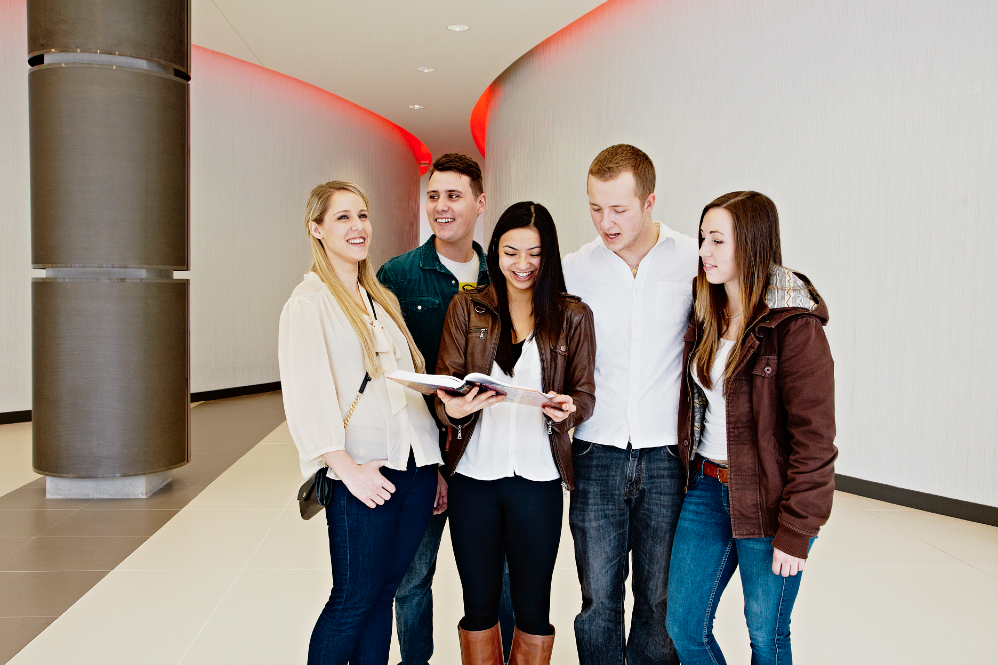 Five students standing and reading a book as a group.