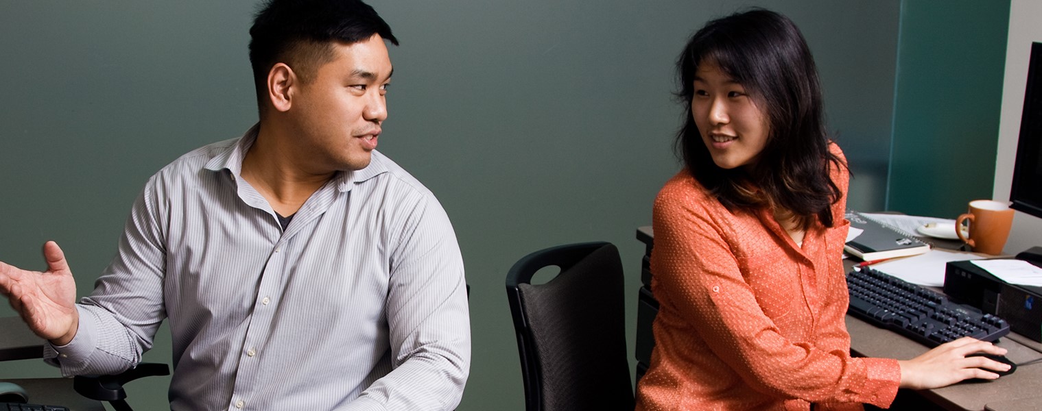 A man and women sitting in front of computers, talking