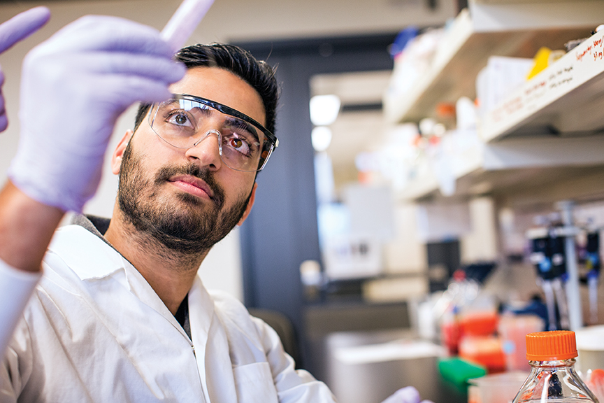 A young man wearing a lab coat, latex gloves, and eye protection working in a lab..