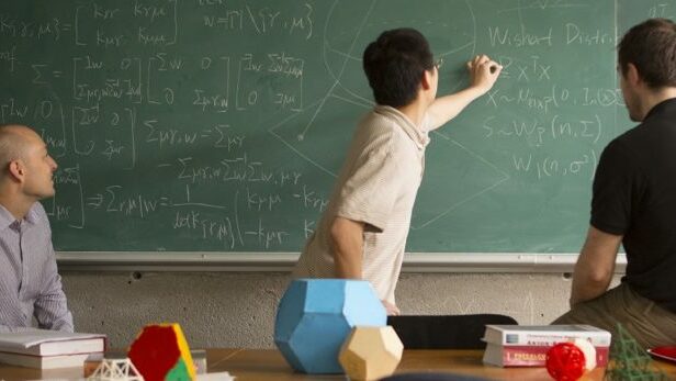 Three men writing on a equations on a green board.