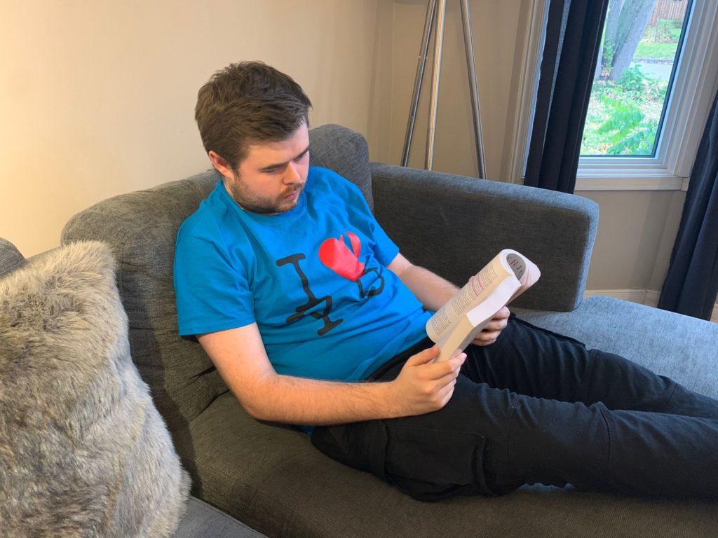 A Learning Skills Services Peer, wearing their Blue uniform shirt, sits on the couch at home reading a book