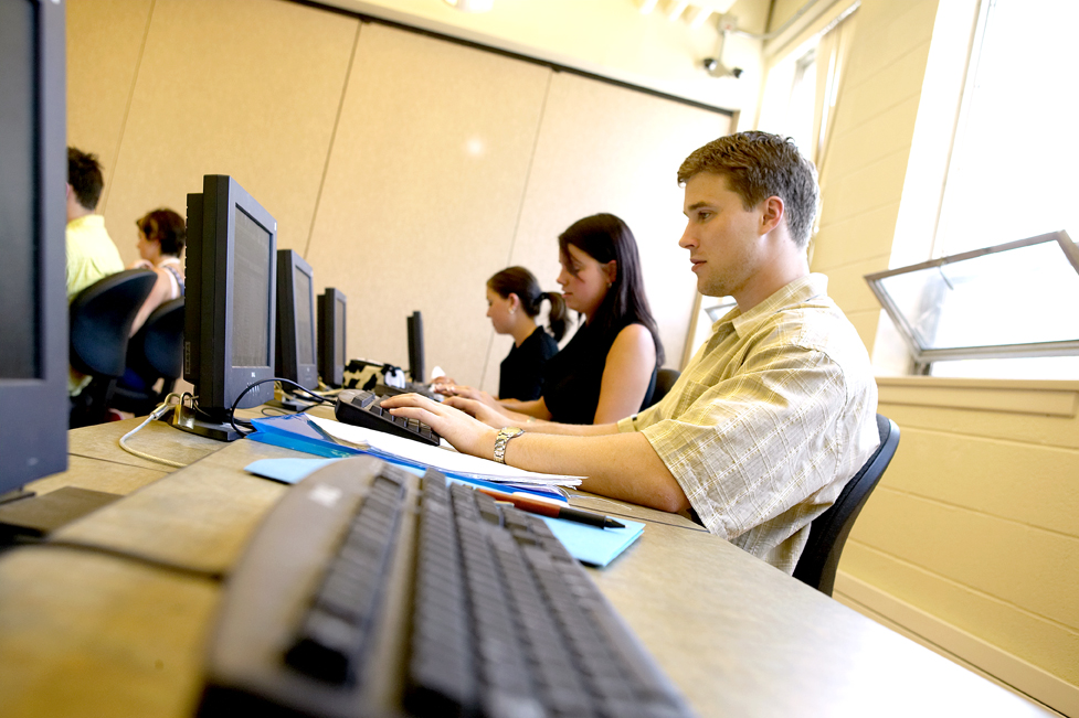 Students sit inside a computer lab typing on computers with note paper and pens on their desks. 