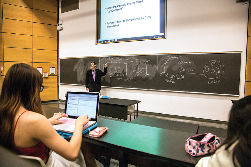 A professor stands at the front of a lecture hall and uses the chalkboard to teach his students. 