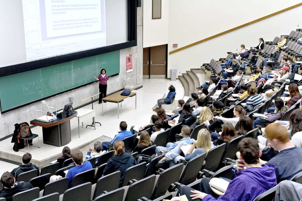 A professor stands in front of a chalkboard in Curtis lecture hall, lecturing a large class of students sitting in their seats. 