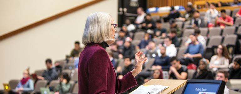 A woman stands in front of a lecture hall, speaking.