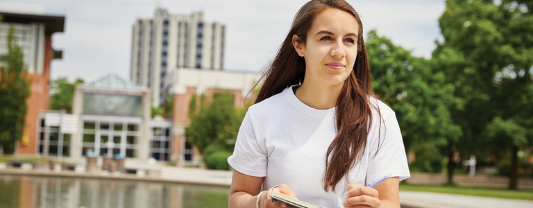 A student sits outside Vari Hall holding a book and pen.