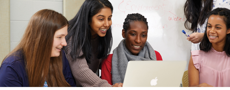 Four students sit together smiling and working together on the same laptop.