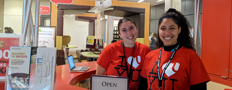 Two Learning Skills Peers stand smiling at the red welcome desk in the Learning Commons, which is located on the second floor of Scott Library.