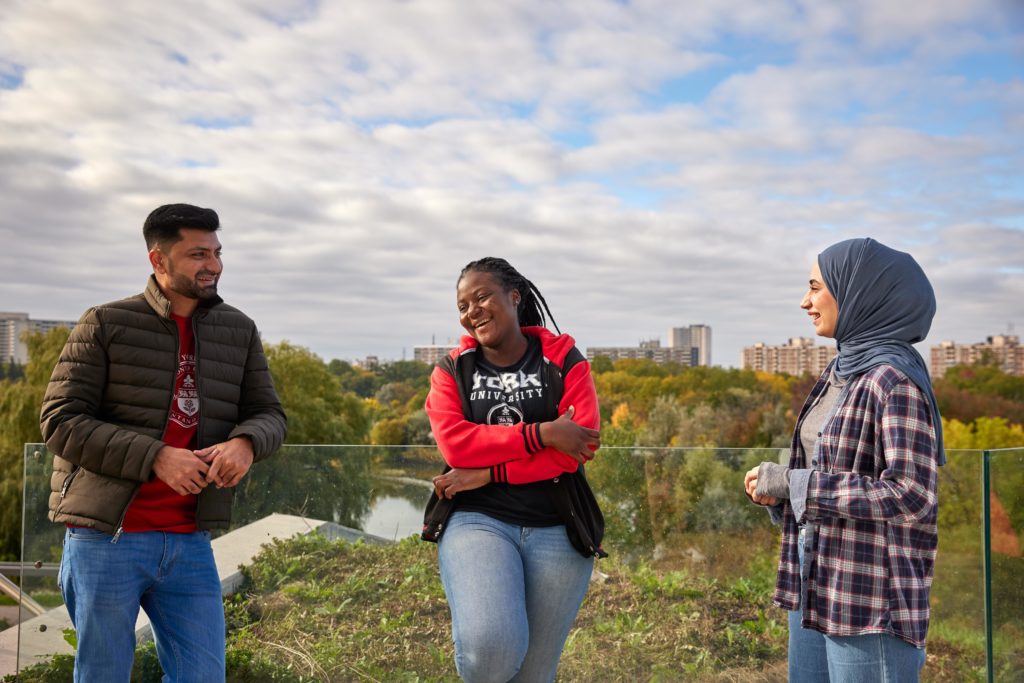 Three York students standing on the balcony of the Bergeron Centre, casually chatting, with a green landscape behind them