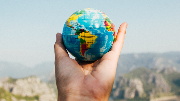 Hand holding a ball with the world map printed on it with South America facing the front. Mountains are blurred in the background.