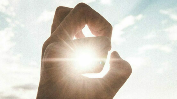 Hand curled in front of sunlight in front of a blue cloud filled sky