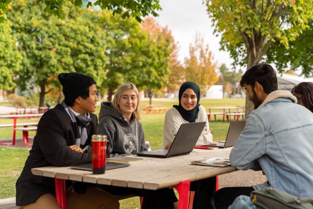 A group of people sitting around a picnic table outside with laptops and paper