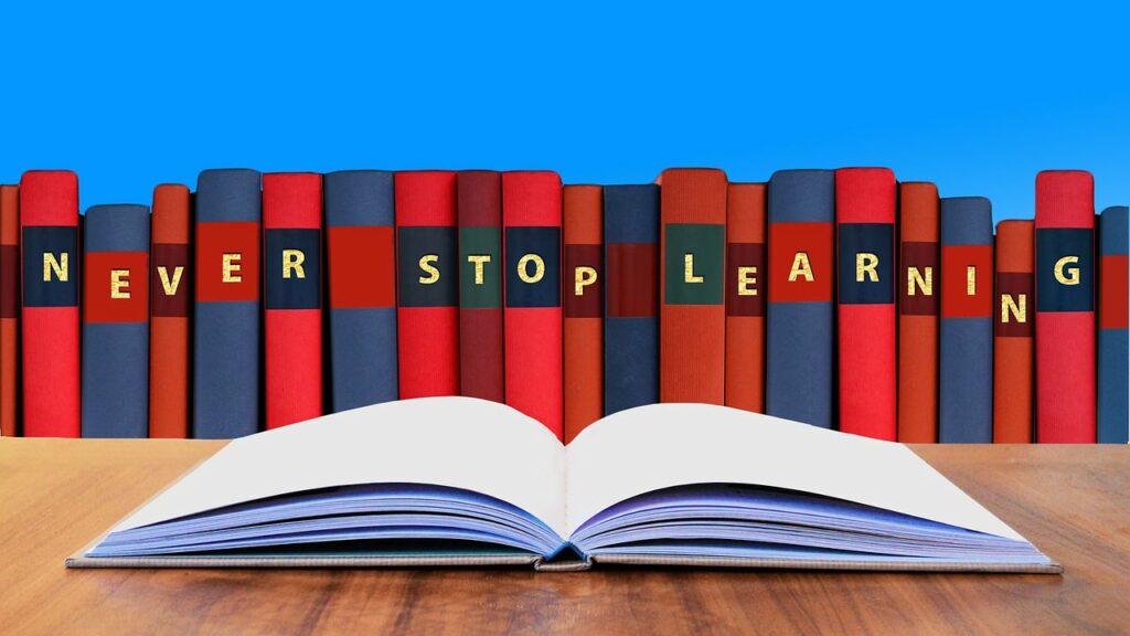 Illustration showing a row of colourful book spines with individual letters on each that spell out 'Never Stop Learning', in front of an open book lying on a table.
