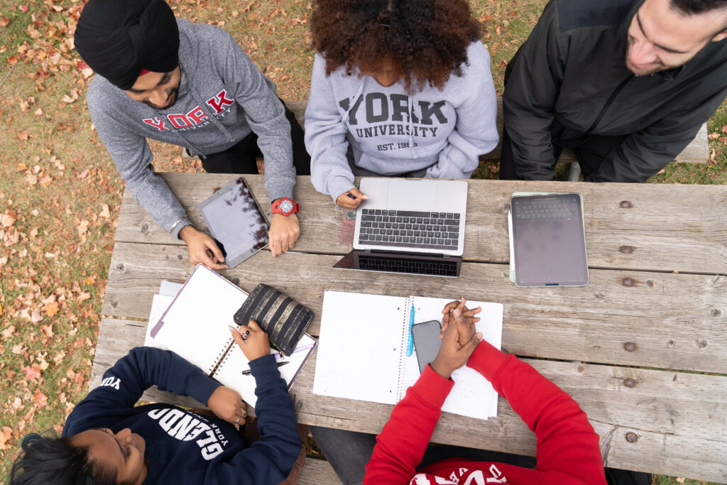 Aerial view of diverse individuals working together at a picnic table with various devices including laptop, tablet, smartphone, and paper and pen.