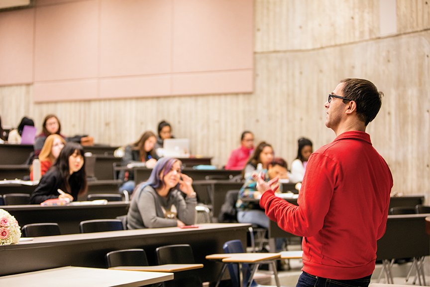 instructor at front of lecture hall teaching to engaged students, image taken from side perspective from behind instructor facing toward group of students
