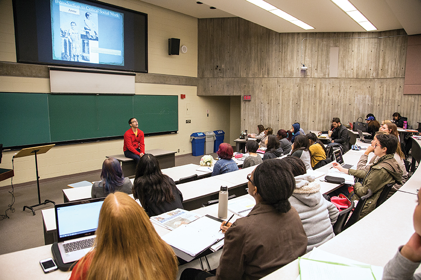 relaxed instructor sitting on desk at front of lecture hall full of engaged students