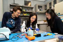 this is a decorative image that shows 3 students sitting a table in a lab room and are wearing safety glasses and looking at equipment on the table