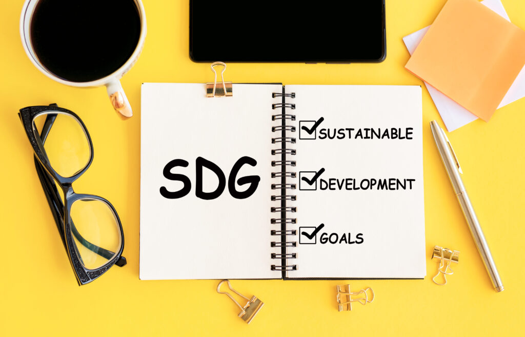 this is a decorative image that has a yellow background with a white notebook and written in black are the words SDG and sustainable and development and goals with check marks next to each word and there is a pair of black eyeglasses with a coffee cup on the left side