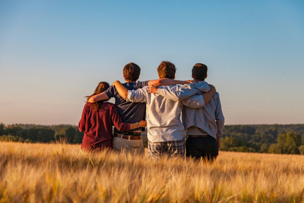 This image shows the backsides of 4 people with their arms around each others standing in a field of brown tall grass. It is a decorative image that represents experiential education