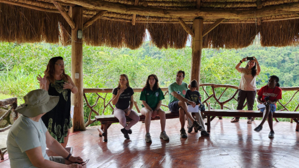 A group of adults sit within a space in the rainforest while one lady is standing and speaking to the group