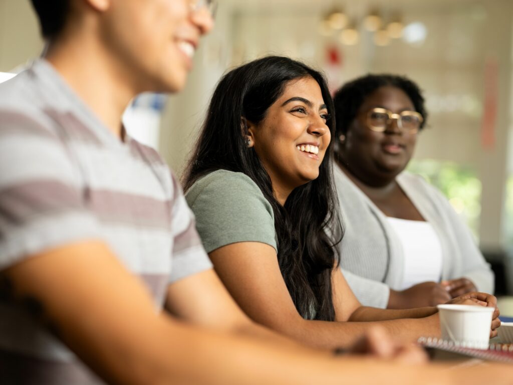 Three students sitting and paying attention to an event, with focus on the student in the middle who has a big smile.