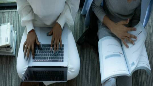 Two black women sitting beside each other with one flipping through a book and another typing on a laptop.