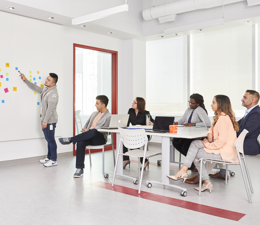 Man standing at whiteboard with five five people sitting listening. 