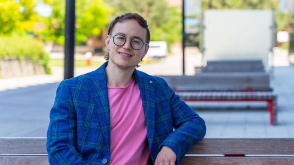Kinnon MacKinnon wearing a pink shirt with a blue blazer and glasses. They have fair skin and short light brown hair. They are sitting on a bench at York's Keele Campus