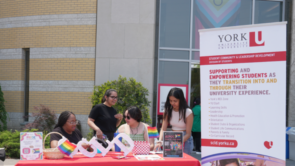 4 york staff stand and sit behind a table with a red table cloth with Pride themed items on top of the table and white SCLd block letters. There are standing banners from York U Alumni and SCLD on each side of the table. 