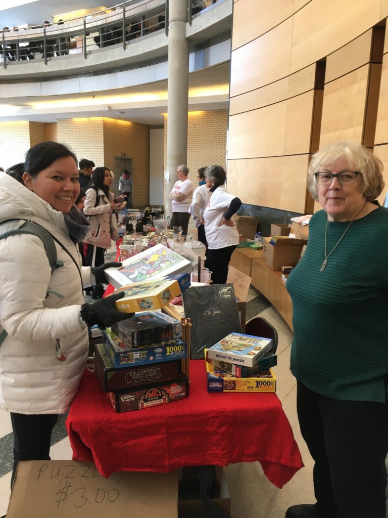 Image of a student holding jigsaw puzzles and a YURA volunteer at the Showcase "attic treasures" table in the Vari Hall Rotunda (with additional volunteers in YURA t-shirts and student customers in the background).