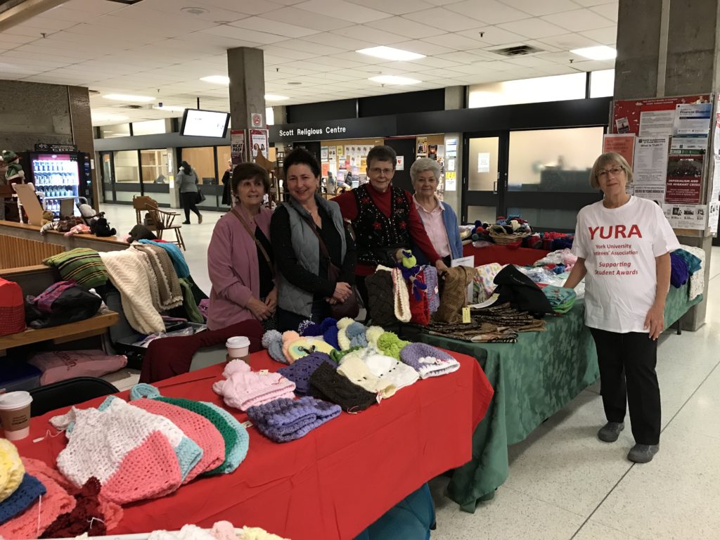 Photo of 4 exhibitors of handmade (knitted, sewn and crocheted) items with a YURA volunteer wearing a YURA t-shirt, standing at the Showcase exhibitor tables.